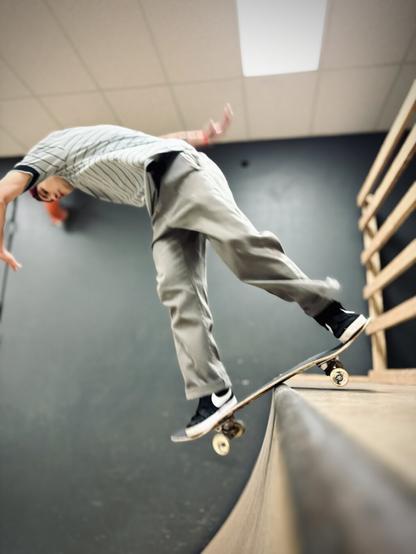 A male skateboarder in a red backwards hat, blue and gray striped polo shirt, gray pants, and black shoes performs a backside disaster on the coping of a larger indoor quarter pipe.
