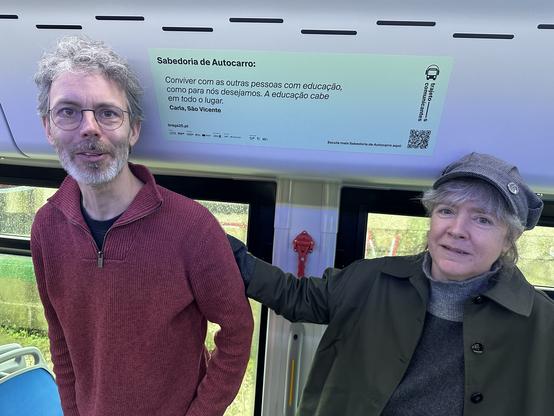 Sarah and Knut stand inside one of Braga's electric passenger buses, inbetween them a sticker that spells out advice for life in Portuguese that was recorded at a bus stop.