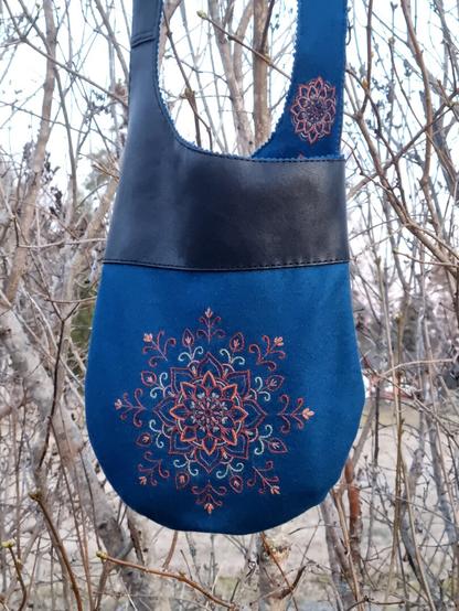 The tote bag hanging in a lilac hedge without leaves. The tote bag is petroleum colored with black leather upper part and belt. On the petrolium colored wool a mandala flower is embroideried in the color of orange, umbra and green.