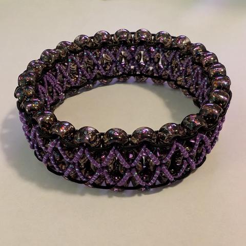 A three row kandi cuff made of black transparent pony beads with rainbow sparkle inside, stitched together using a ladder stitch of black elastic on the outer rows and purple elastic woven with opalescent filament in a diamond/X pattern holding the rows together.