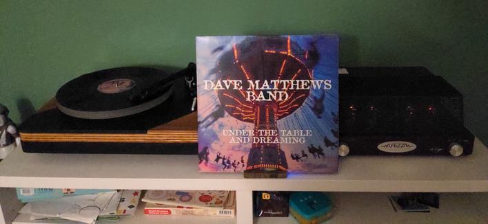 Giradischi spinna un vinile nero. Alla sua sinistra la copertina del disco ed un amplificatore.