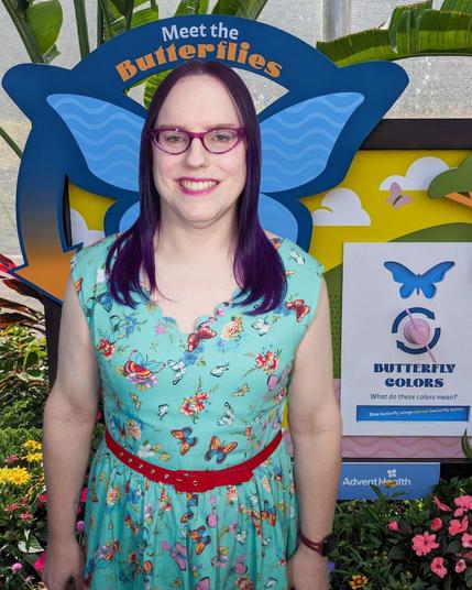 The image features a woman standing in front of a colorful backdrop, likely at an event or exhibit related to butterflies.

In the foreground, the woman is dressed in a turquoise dress adorned with pink and orange flowers, complemented by a red belt around her waist. Her dark hair cascades down her back, framing her face as she gazes directly at the camera with a warm smile. The woman's attire and demeanor suggest that she is an attendee or staff member at the event.

Behind the woman, a vibrant backdrop serves as the primary focus of the image. A large blue butterfly silhouette dominates the center, accompanied by the words "Meet the Butterflies" in white text above it. To the right of the butterfly, a smaller logo featuring a blue butterfly and the phrase "Butterfly Colors" is prominently displayed.

The overall atmosphere of the image exudes excitement and curiosity, as if the woman is eager to explore the wonders of butterflies. The colorful backdrop and playful graphics contribute