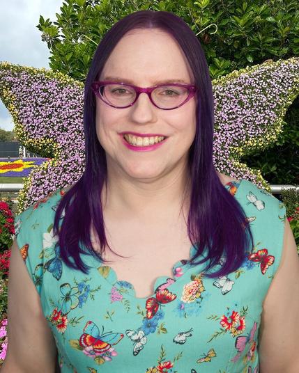 The image features a woman standing outdoors, facing the camera directly. She has long dark hair that falls just below her shoulders, and is wearing a turquoise top adorned with vibrant floral patterns in shades of red, blue, yellow, pink, and white. The woman's attire also includes a pair of glasses with purple frames.

In the background, a lush green hedge or bush stretches across the entire width of the image, punctuated by small clusters of purple flowers that add a pop of color to the scene. Above the hedge, a sliver of blue sky is visible in the top left corner, providing a subtle hint at the surrounding environment.