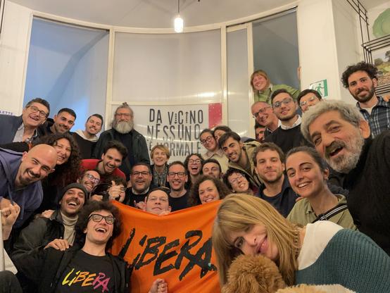 Foto di gruppo mostrando la bandiera di Libera. Dietro, scritta sul muro “da vicino nessuno è normale”