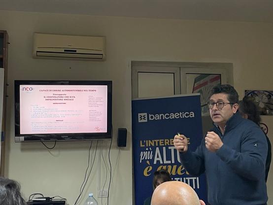 Peppe che racconta la storia di NCO. Dietro il banner di Banca Etica: ”L’interesse più alto è quello di TUTTI”