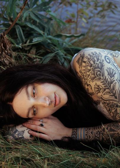 A tattooed woman laying on grass in front of a lake