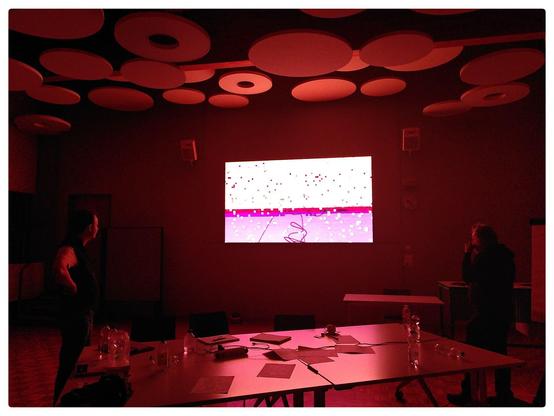 People staring at a big LCD screen, showing Markov Chant - a PC Demo released at MountainBytes 2025