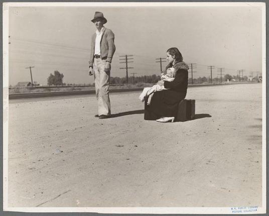 The image is a black and white photograph depicting two individuals on what appears to be an open dirt road. To the left, there's a man wearing casual attire including jeans, boots, and a jacket over his shoulders along with sunglasses atop his head as he walks away from the camera with his right hand in his pocket and looking down towards the ground. On the opposite side sits a woman dressed in dark clothing holding an infant on her lap while resting against what seems to be a suitcase or similar container beside her. She is smiling slightly, presumably at something outside of the frame.

In the background lies flat terrain with sparse vegetation and telephone poles extending across the horizon under a clear sky. The overall atmosphere suggests economic hardship during hard times like depression era America where such images were taken as part of documentation for social studies or journalistic purposes to portray real-life stories affecting people's lives at that time in California, USA.

Further details on this particular image are found through provided references which indicate its origin from the NY Public Library Picture Collection and a specific mention by Young family. It was created early during 1935 showcasing hardships faced like being penniless while hitchhiking along U.S. Highway 99 with background information stating they came from Winston-Salem, North Carolina before this time period which gives context t [...]