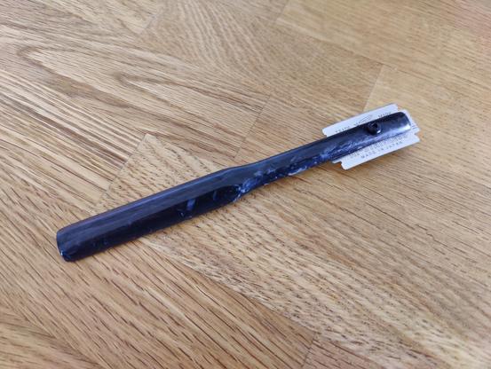 A baker's lame made of forged carbon. The handle is a very simple shape, like a half cylinder. The safety razor blade is slightly curved and held onto the handle with a screw.