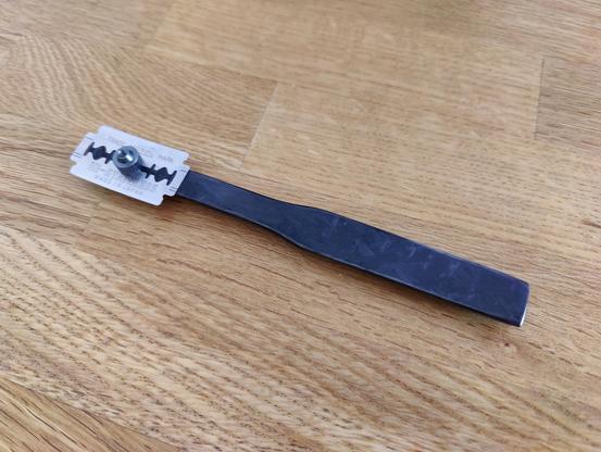 A baker's lame made of forged carbon. The handle is a very simple shape, like a half cylinder with narrower neck on one side. The safety razor blade is slightly curved and held onto the handle with a thumb-nut.