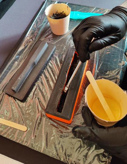 The moulding process which involves drenching some chopped carbon tow together with infusion epoxy resin and packing the material inside the cavity of a 3D-printed mould. The compression half of the mould is visible next to it.