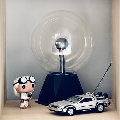 A Pop! figurine of Doc Emmett Brown, holding a RC remote control, looking towards a scale model of the Delorean from the movie Back To The Future. There is also a large plasma ball in center frame. All exists with a neutral grey background within an IKEA shelf square.
