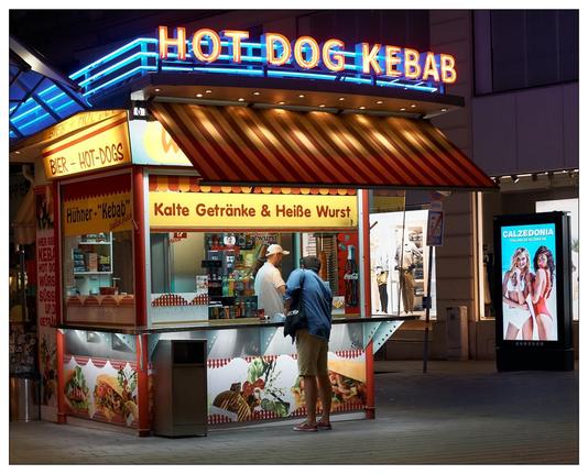 This image shows a street food kiosk at night, brightly lit and featuring neon signs that advertise "HOT DOG" and "KEBAB." The kiosk sells a variety of items including beer, hot dogs, kebabs, and both cold drinks and hot sausages, as indicated by the signage. There is a customer at the counter engaging with the vendor who is behind the counter. Additionally, there's an illuminated advertising display to the right showing an advertisement for Calzedonia, a fashion brand. The setting appears to be urban, and the overall atmosphere conveys a casual, evening snack spot vibe.