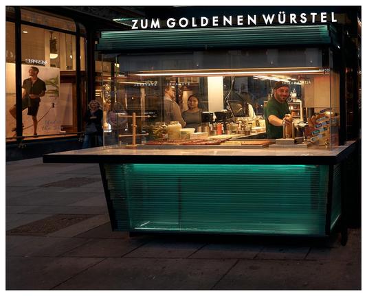 This image depicts a vibrant street food vendor stall named "Zum Goldenen Würstel," located in an urban environment during the evening. The stand features bright lighting and a clean, modern design with a luminous turquoise-colored front. Inside the stall, a man who appears to be preparing food for two others on the other side of the glassed in booth.  The man has spotted the camera and is smiling directly at us.  Outside, there are pedestrians passing by. The setting conveys a lively city atmosphere with a focus on street food culture.