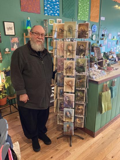 Photograph of a balding man with a beard standing by a greeting card rack featuring his artwork.