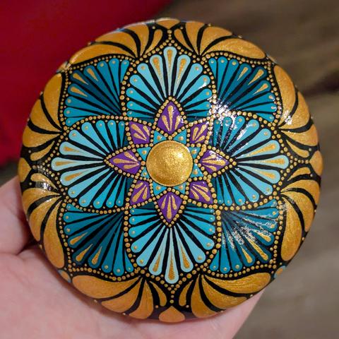 A photo of a round, flattish dot painted art stone. The main geometric design is composed of flower-like petals radiating around a star shape in the centre. The star has a large gold dot in the centre and eight purple "points". The petals around this star are filled with radiating teardrop shapes in gradient shades of turquoise and accented with gold. It's all painted on a black background.
