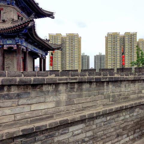 Part of the fortification wall, an old-style sentry building to the left, two red lanterns on poles, and residential highrises in the background.