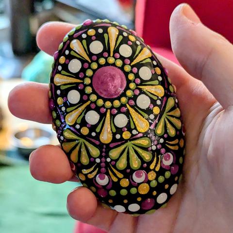 A small oval-shaped dot painted art stone, held loosely in my hand. The design is a rather simple or "classic" dot painted design, with a large centre dot and radiating circles of dots and teardrop designs surrounding it in colours of metallic pink, pale apple green, white, and metallic gold painted on a black background.