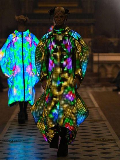 Two models walk the runway in dramatic, multicolored LED-lit garments and towering platform shoes.