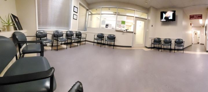 A brightly lit waiting room features multiple rows of blue, armless chairs arranged neatly. A reception window is present at the end of the room, with signage visible. Further into the space, a sign indicating "Staff Only" is posted above a hallway, suggesting a restricted area. A television is wall-mounted above a row of seats. The floor is grey. The room emphasizes cleanliness and order.