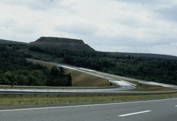 A four lane divided highway curving in a reverse C. At the end of the curve above the line of trees is a mountain or hill that has been flattened probably by mining.