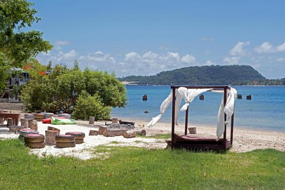 Photo shows a view to a harbour with deep blue green water with an island on the other side. The sky is a kobolt blue  with some light clouds in the horizon. In the foreground is a collection of furniture for sun lowers, including a sun bed with lampposts with light white covers. They are placed in the changing zone between the green turf and the white sand of the beach. The beach stretches out behind the furniture down to the water.