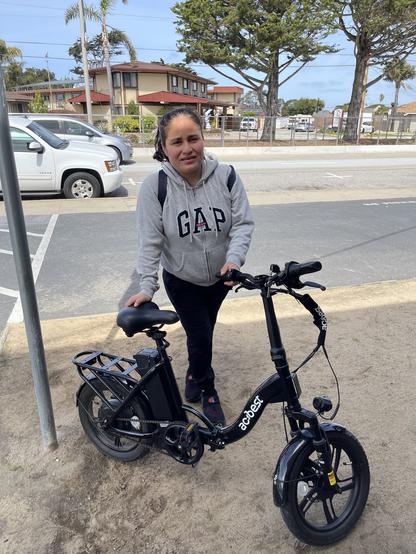 A young woman, her black hair tied back in a ponytail, is dressed in a grey Gap hoodie, black pants, and black shoes. She is standing alongside her black Actbest CityBike, a low-to-the-ground e-bike.