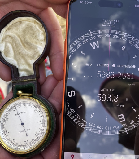 On the left, the camera is looking down at a pink hand holding a small brass pocket barometer/altimeter in a small purple antique case. The needle on the dial is indicating a height of 600 feet. On the left, a different hand is holding a modern digital GPS which displays an altitude of "593.8 +- 14 feet".