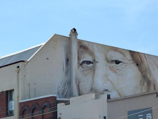 The image features a large mural painted on the side of a building. The mural depicts a close-up of an elderly person's face, with detailed features such as wrinkles, eyes, and hair. The face is rendered in shades of beige and white, creating a realistic and textured appearance. The mural covers a significant portion of the building's facade, extending from the top to the middle of the structure. The building itself has a white brick exterior with a metal roof, and there is a chimney visible on the left side of the mural. Below the mural, there are other buildings with different architectural styles, including a red brick building with arched windows. The sky is clear and blue, indicating a sunny day.
