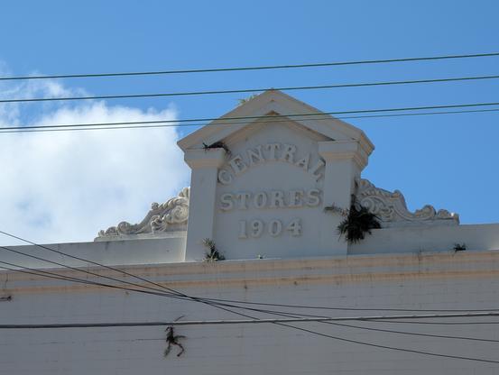 The image shows the top portion of a white building with the inscription "CENTRAL STORES 1904" prominently displayed in raised letters on a decorative pediment. The building's facade is plain, with a clear blue sky in the background. Power lines are visible in the foreground, crossing in front of the building. The pediment features ornate carvings, adding a touch of architectural detail. The building appears to be in good condition, with some minor signs of wear, such as small plants growing on the edges of the structure. The overall scene is bright and sunny, with the building standing out against the clear sky.