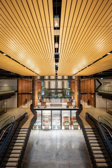 Interior of Battersea Power Station, featuring a grand atrium with a modern industrial design. A large central staircase with black railings and wooden steps leads down to a spacious open area with polished concrete flooring. Above, a wooden slatted ceiling with hanging rectangular lights creates a warm ambiance. The walls feature exposed red brick, large glass panels, and black metal accents. In the background, a mezzanine level with greenery, seating areas, and glass railings is visible.