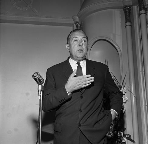 Black and white photo of Jacques Tati in a dark suit, speaking behind a microphone. He is making a hand gesture, has a lot to explain probably.