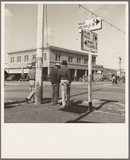 The image is a black and white photograph depicting two men standing at an intersection, looking in the direction of various street signs. The man on the left wears a wide-brimmed hat and jeans, while the other sports a cowboy-style hat and dark pants. They appear to be engaged in conversation as they glance toward one sign pointing towards "West Holtville," with distances mentioned alongside: 9 miles for West Holtville; 20 miles for El Centro; Niland Medca is noted on another post nearby.

Behind them, there's a bustling street scene including various vehicles and pedestrians. A large building stands in the background which could be indicative of local businesses or shops typical to that era. The setting suggests an old western town with its architecture style reminiscent of mid-20th-century America.

The photograph exudes a sense of nostalgia, possibly from California due to contextual clues like "Holtville" and "Niland Medca." It captures the essence of rural life during this period, highlighting aspects such as employment prospects for those in agricultural sectors.