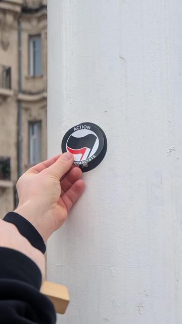 Photo d'une main qui recouvre le sticker croix celtique d'un sticker action antifasciste anarchiste, sur un poteau blanc.
