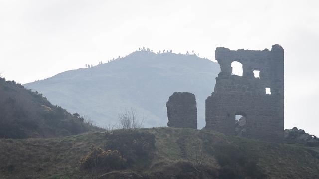 A photo taken in Holyrood Park, in Edinburgh, looking up past a ruined St Anthony's Chapel to the distant silhouetted hilltop of Arthur's Seat behind, absolutely covered with people. Everything is pale and hazy.