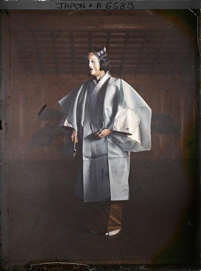 A tinted photograph of a single Noh performer on what appears to be a traditional wooden stage. The performer wears a pale, layered garment with wide sleeves, resembling a formal kimono ensemble, and a white mask with a calm expression. The mask covers the entire face, leaving only a slight gap around the jawline. The outfit’s upper layer is a light gray or blue robe with subtle decorative patterns, extending to knee length. The sleeves and collar display white embroidered details, while the performer’s lower garment seems to be a darker hue, partially visible near the feet. Traditional footwear, likely tabi socks and sandals, can be seen. A slim, possibly ceremonial fan or small instrument is held in one hand near the waist. The performer’s hair is arranged in a stylized manner or is part of the mask, with a small ornamental piece on top. The background shows wooden walls and a ceiling typical of a Noh theater, with muted tones that frame the performer. The words “JAPON A 6589” are written at the top edge of the photograph. Some discoloration and dark spots appear around the border, suggesting age or damage to the plate. The overall scene conveys a moment from a classical Japanese Noh performance, emphasizing the refined costume, mask, and stage setting.

Photo by Stéphane Passet, CC0, via Wikimedia Commons