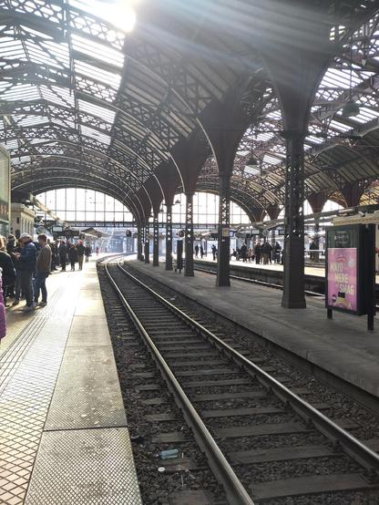 View from a train platform down the tracks towards a sunny outside. There are arches overhead and people standing on the platform. The track gradually bends round to the right out of sight