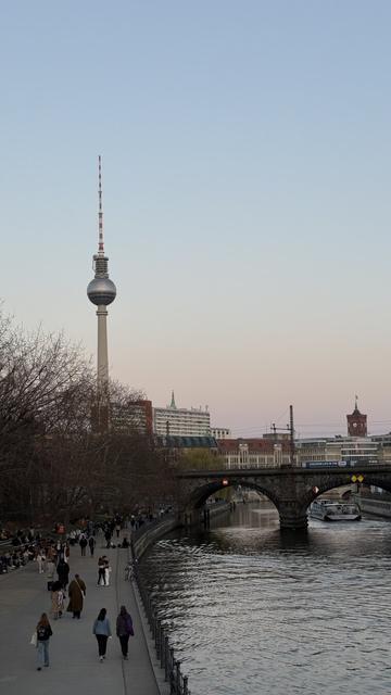 La imagen muestra una vista urbana al atardecer en Berlín, Alemania. En el centro se alza la icónica Torre de Televisión (Fernsehturm), con su característica esfera metálica y antena roja y blanca. A la derecha se ve un puente de piedra cruzando el río Spree, sobre el cual pasa una línea de tren. En el agua navega un barco turístico, mientras que a la izquierda, junto al río, muchas personas caminan o se sientan en bancos disfrutando del paseo. La escena transmite una atmósfera tranquila, con el cielo despejado en tonos pastel que anuncian el final del día.