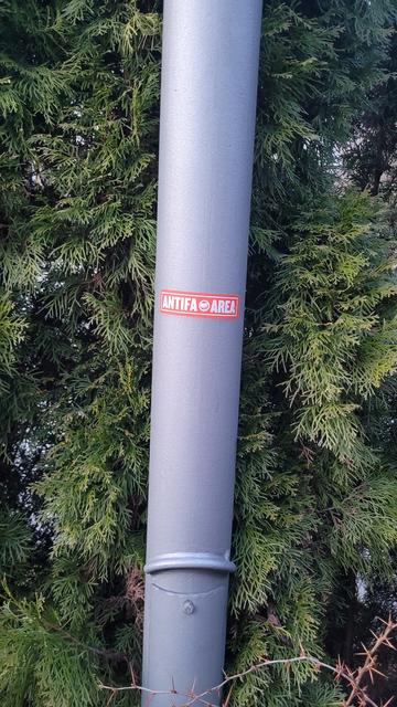 Ein grauer Laternenpfahl steht vor einer grünen Thujahecke.
Am Pfahl klebt ein rechteckiger, roter Aufkleber,  auf dem in weißer Schrift 'Antifa Area' steht. Zwischen den beiden Worten sind die Antifafahnen abgebildet.