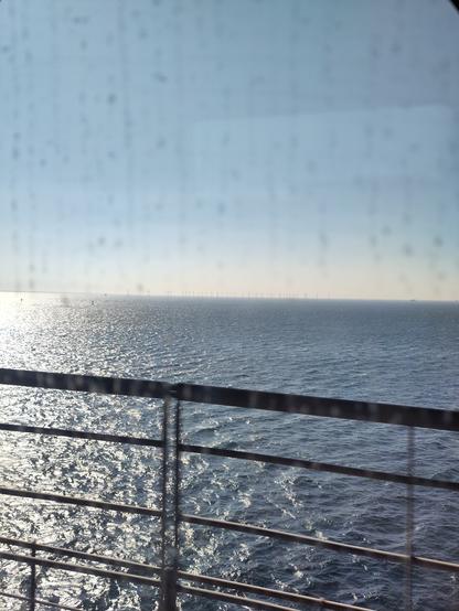 Blurry view from a train window of blue sea, and a bridge handrail. In the far distance wind turbines under a blue sky.