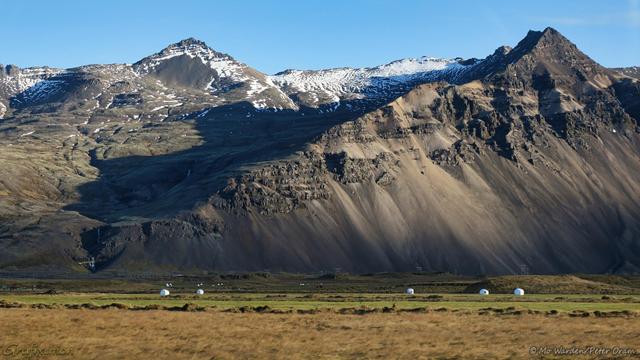A colour photo of two in a range of mountains, beneath a clear cyan sky. The foreground is yellow vegetation, some fields are still green. The mountain flanks on the right are steep scree slopes, and a shadow is cast between the two peaks, highlighting a deep valley. The pinnacle on the left looks like harder rock, and the peaks here are ice- and snow-capped.