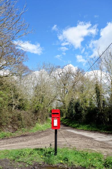A vibrant red Royal Mail post box stands centrally on a black metal post beside a rural dirt road. The post box has a white collection time notice visible on its front. The road stretches into the distance, flanked by bare trees and green foliage under a bright blue sky dotted with fluffy white clouds.