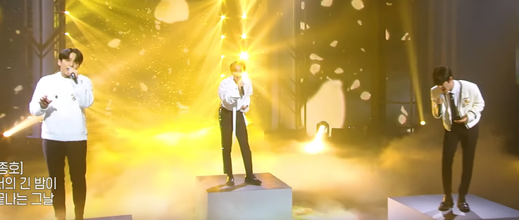 Jongho, Eunkwang and Seungmin standing on individual plinths among dry ice fog with golden lights shining from behind them