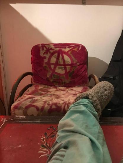 photo of comfortable arm chair that has been tagged with anarchist A with queer symbols, also my leg with a fluffy shoe on