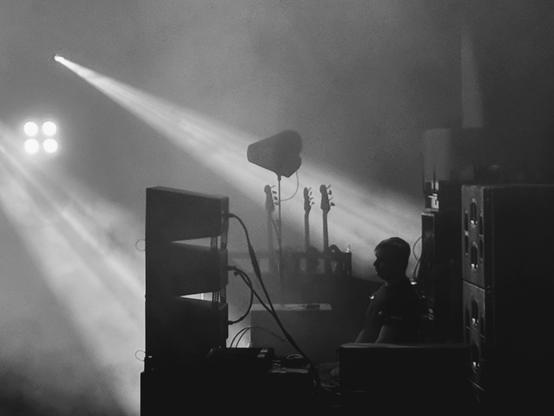 A concert technician sits patiently in the shadows on the side of the stage during a rock performance, with atmospheric smoke and lights surrounding them.