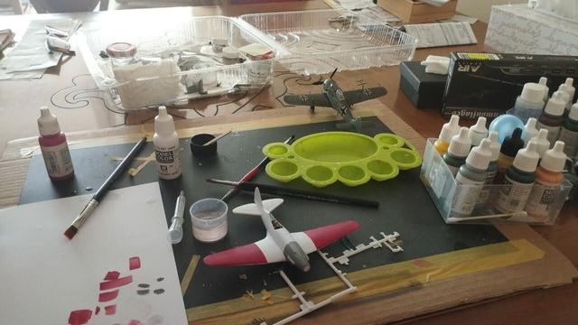 Mesa con herramientas y pinturas de modelismo. En primer plano hay un Mig-3 a medio montar pintado en blqnco y rojo. Al fondo puefe verse un FockeWulf 190