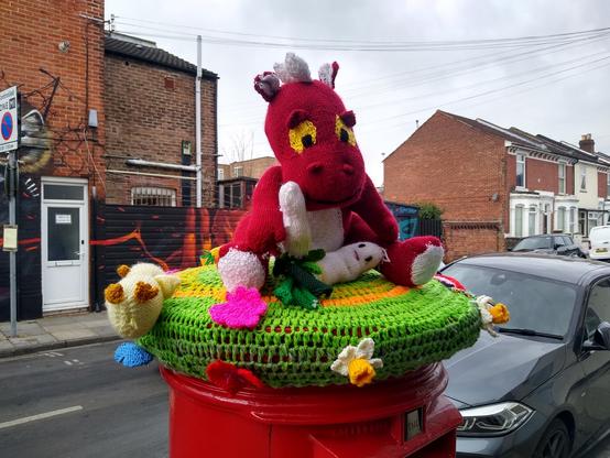 Photograph of a knitted post box topper, with a large red dragon and a leek with a face on its lap, daffodils around the edge and a chick