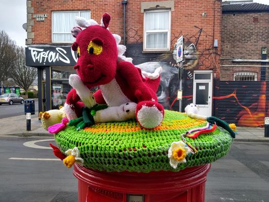 Another photograph of the knitted postbox topper with the large red dragon, a peek on its lap and daffodils