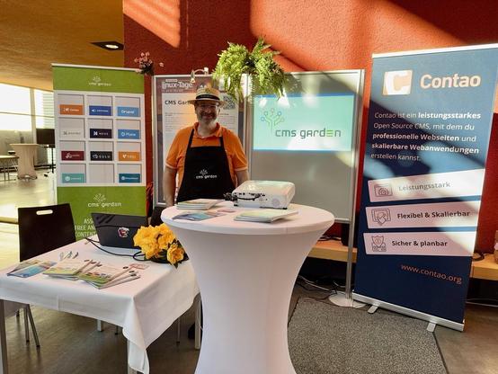 Joachim mit markantem CMS Garden Hut und Schürze auf der bunten Standfläche des CMS Garden. Links und rechts zwei CMS-Rollups. mittig eine Projektion per Beamer.