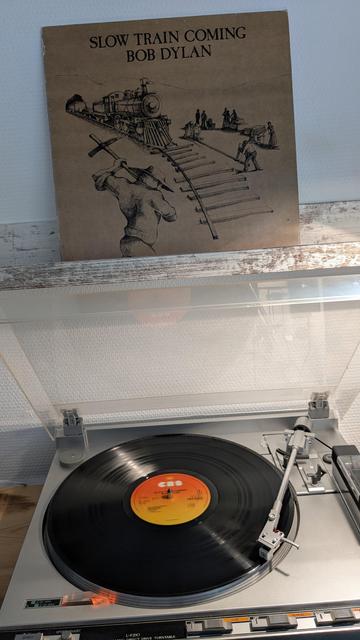 Das Bild zeigt einen Plattenspieler mit einer Vinyl-Schallplatte, die gerade abgespielt wird. Die Schallplatte hat ein orangefarbenes Label mit dem CBS-Logo. Über dem Plattenspieler steht das Albumcover von Bob Dylans "Slow Train Coming". Das Cover ist in einem schlichten, sepiafarbenen Design gehalten und zeigt eine Zeichnung eines Zuges, der auf Schienen fährt, während Arbeiter die Gleise verlegen. Im Vordergrund ist ein Mann mit einem Werkzeug zu sehen. Der Plattenspieler hat eine silberne Oberfläche und ist mit einer durchsichtigen Abdeckung ausgestattet.